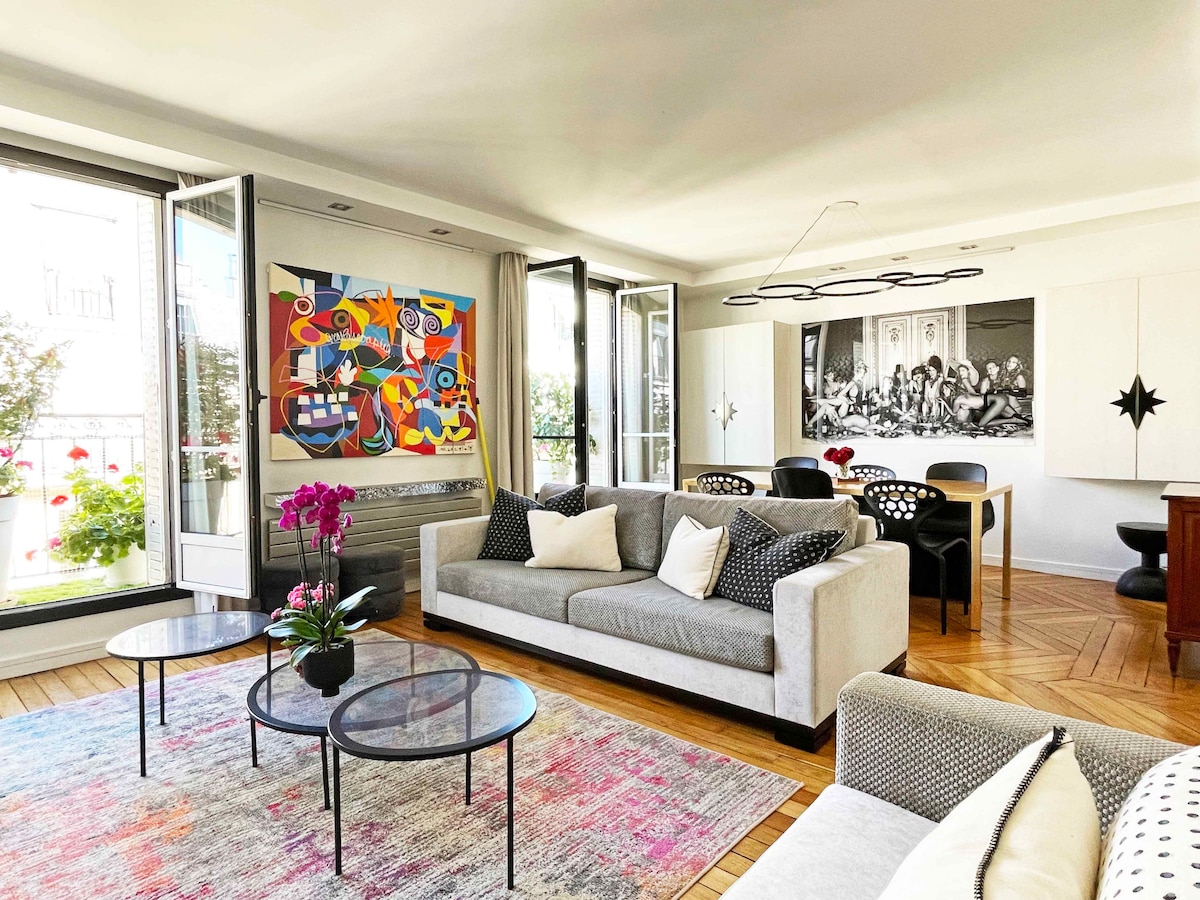 The living area features a mix of contemporary furniture and vibrant artwork. A light grey sofa is complemented by patterned cushions. Two round glass tables are positioned over a colorful area rug, while large windows allow natural light to fill the room, framed by floral balconies.