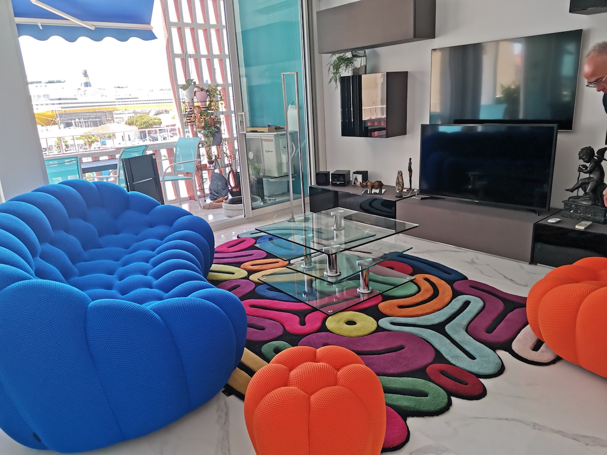 A vibrant living area features a blue, tufted chair and orange poufs arranged on a colorful rug. A glass coffee table sits atop a marble floor, complemented by a wall-mounted TV and decorative elements. Large windows allow natural light to fill the space, providing views of the port.
