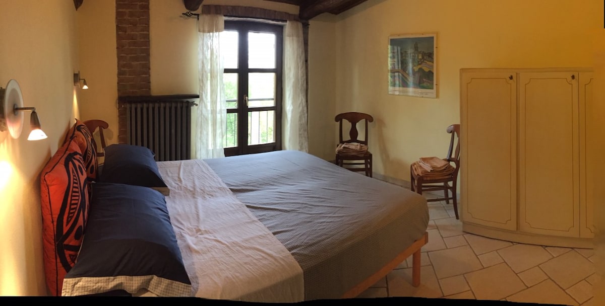A light-filled room features a spacious bed with dark blue and patterned bedding. A wooden cabinet and two chairs complement the warm yellow walls. Natural light pours in through the large window, framed by sheer curtains, illuminating the rustic brick walls and tiled floor.