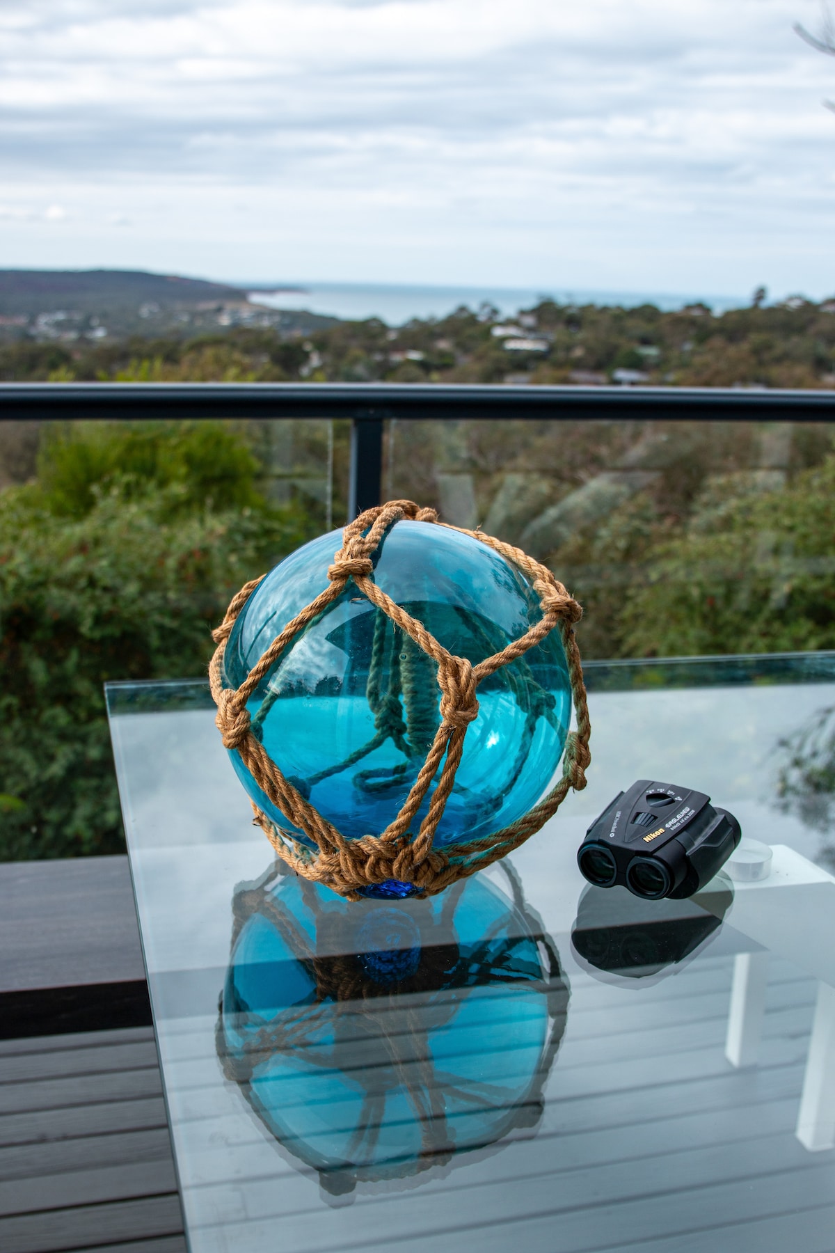 A glass buoy wrapped in decorative rope is placed on a table, reflecting the surrounding greenery and coastal view. A pair of binoculars sits nearby, suggesting opportunities for observation and exploration of the scenic landscape.
