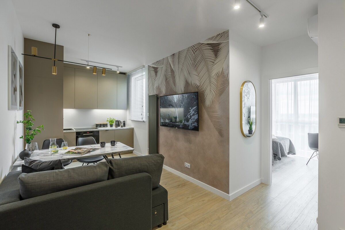 A modern living area features a dark sectional sofa facing a wall-mounted television. A dining table is set with decorative items, and the open kitchen displays sleek cabinetry. Natural light floods through large windows, creating a bright and airy ambiance.