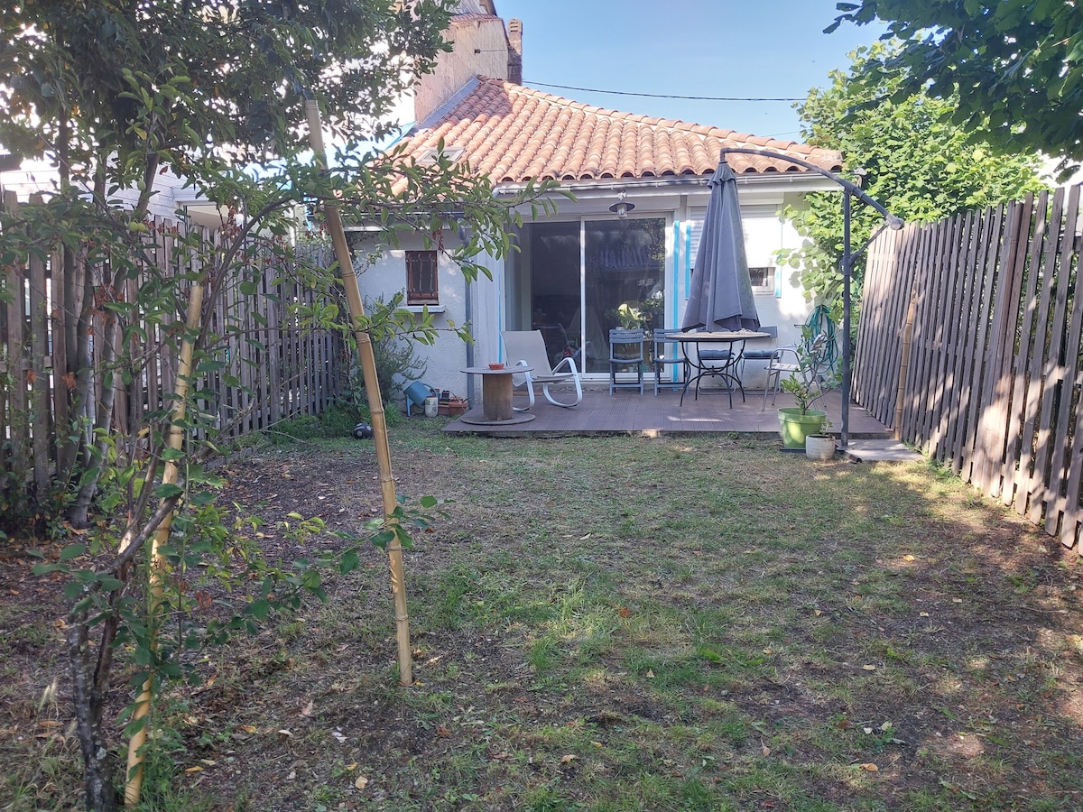 A serene garden area is visible, featuring a wooden terrace with a table and chairs beneath an umbrella. Lush green grass is present, along with trees and a wooden fence bordering the space. A glimpse of the home’s exterior is seen in the background.