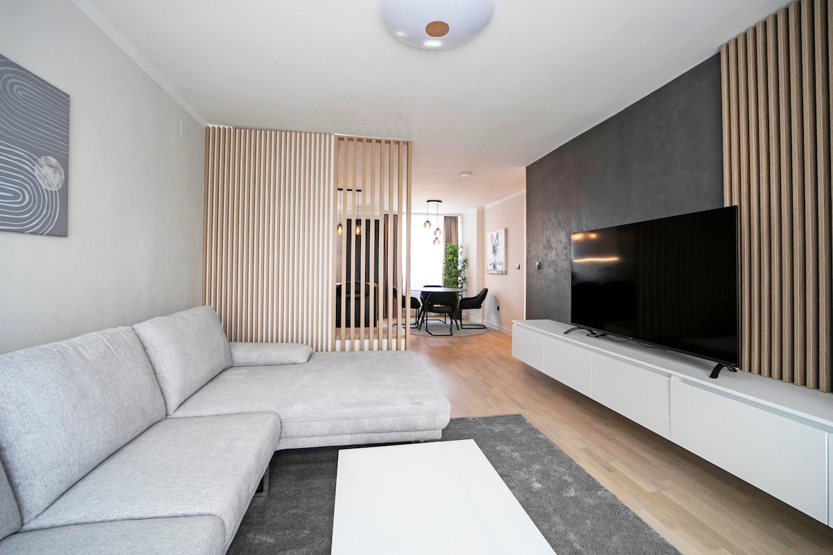 A modern living area features a sleek sectional sofa and a minimalist coffee table on a soft rug. A large television is mounted on a white media console, while light wood flooring creates a warm ambiance. Partitioning adds a sense of separation to the dining area.