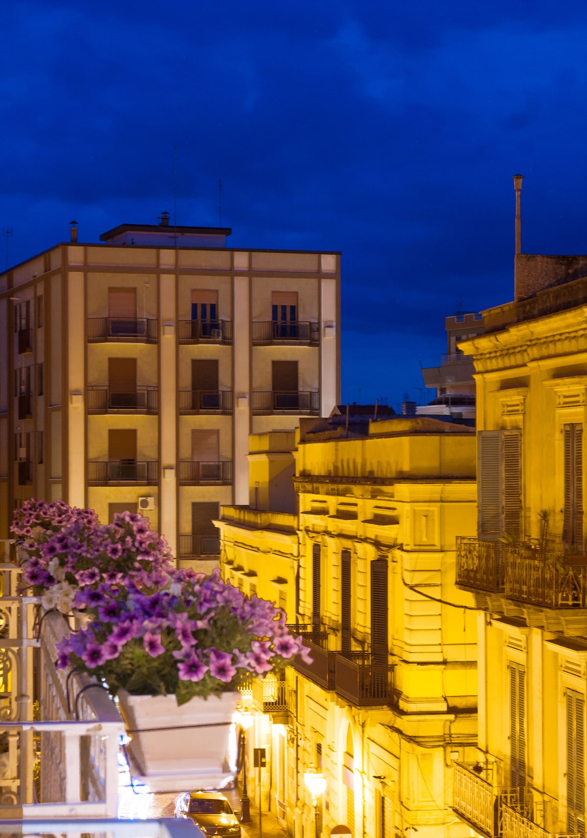 A cityscape is captured at dusk, showcasing buildings with warm golden lighting. Colorful flower pots line a balcony, adding a touch of nature against the architectural backdrop. The sky transitions to a deep blue, enhancing the evening ambiance of the urban scene.