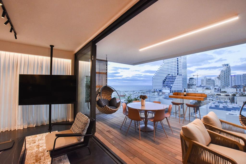 A spacious terrace features a dining area with a round table and several chairs, complemented by a comfortable seating area. Large glass doors open to showcase city and sea views, while hanging chairs add a unique touch to the outdoor atmosphere.