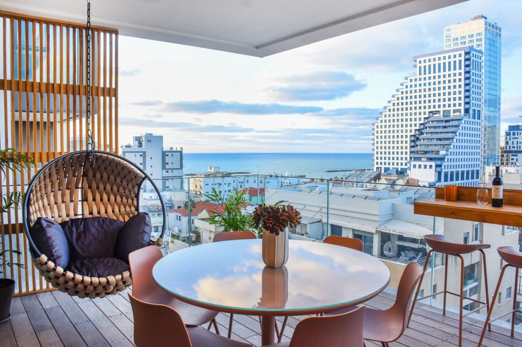 A terrace offers a panoramic view of the Mediterranean Sea and cityscape, featuring a round table surrounded by sleek chairs. A hanging wicker chair adds a cozy spot to relax, while potted plants provide a touch of greenery amidst the modern architectural backdrop.