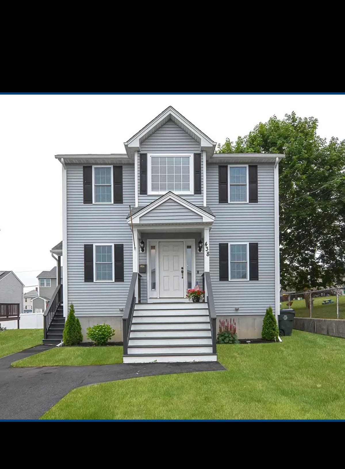 A three-story gray home features a gabled roof and front porch, framed by neatly trimmed bushes. White steps lead to a welcoming entrance, while dark shutters contrast against the siding. The well-maintained lawn provides a sense of openness, with a paved driveway extending from the street.
