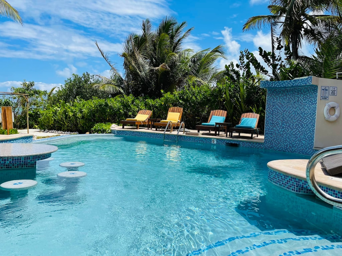 A refreshing resort pool glistens in sunlight, surrounded by tropical greenery. Lounge chairs in vibrant colors are positioned around the water's edge, inviting relaxation. The pool features built-in seating and a shallow area, ideal for lounging and enjoying the tranquil atmosphere.