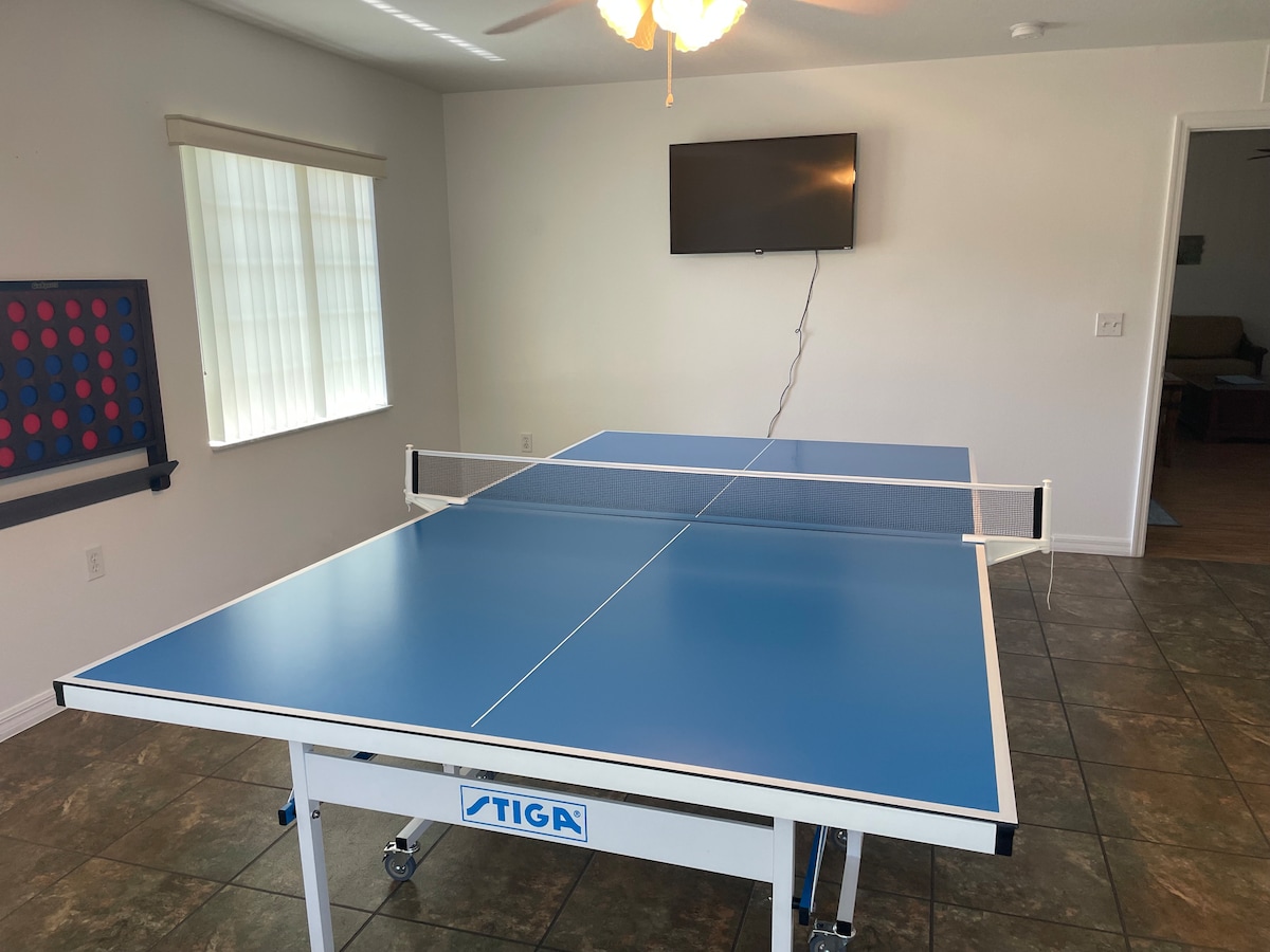 A blue ping pong table is centrally positioned in a well-lit bonus room, accompanied by a wall-mounted TV. Natural light filters through the adjacent window, with blinds partially drawn, contributing to the room's spacious feel. A ceiling fan is visible above, enhancing air circulation.