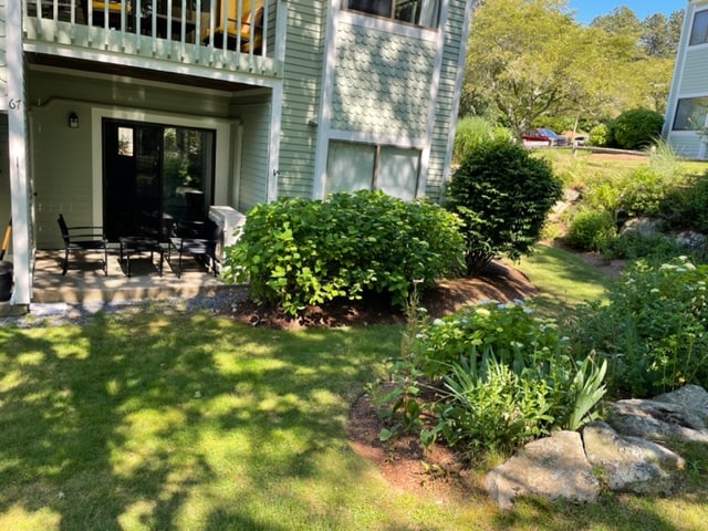 A private patio area is visible, furnished with a black dining set surrounded by lush greenery. Soft sunlight filters through the trees, casting gentle shadows on the neatly manicured lawn. The patio is bordered by clusters of shrubs and flowering plants.