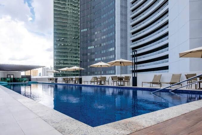 A serene pool area is showcased, featuring clear blue water and surrounded by lounge chairs with umbrellas. The modern architectural skyline provides a contemporary backdrop, while spacious deck areas invite relaxation and leisure.