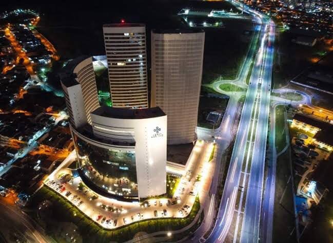 An aerial view showcases a modern building complex illuminated against the night sky. The structure features a sleek design with multiple levels, surrounded by a well-lit roadway and parking area. Nearby, city lights and infrastructure create a vibrant urban atmosphere.