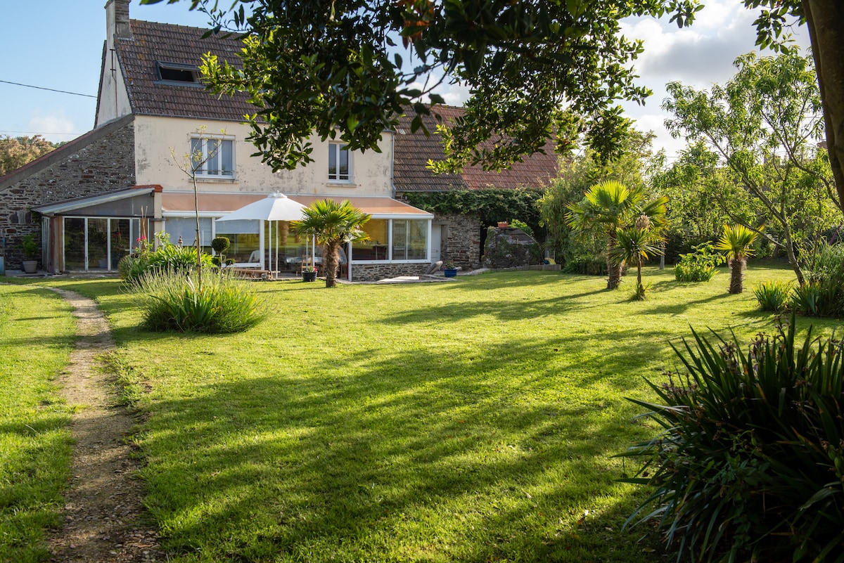 The exterior of the renovated farmhouse is shown, featuring a spacious lawn with well-maintained greenery and palm trees. A shaded seating area is visible under an awning, with large glass windows reflecting the yard. A path leads through the landscaped garden, enhancing the peaceful setting.