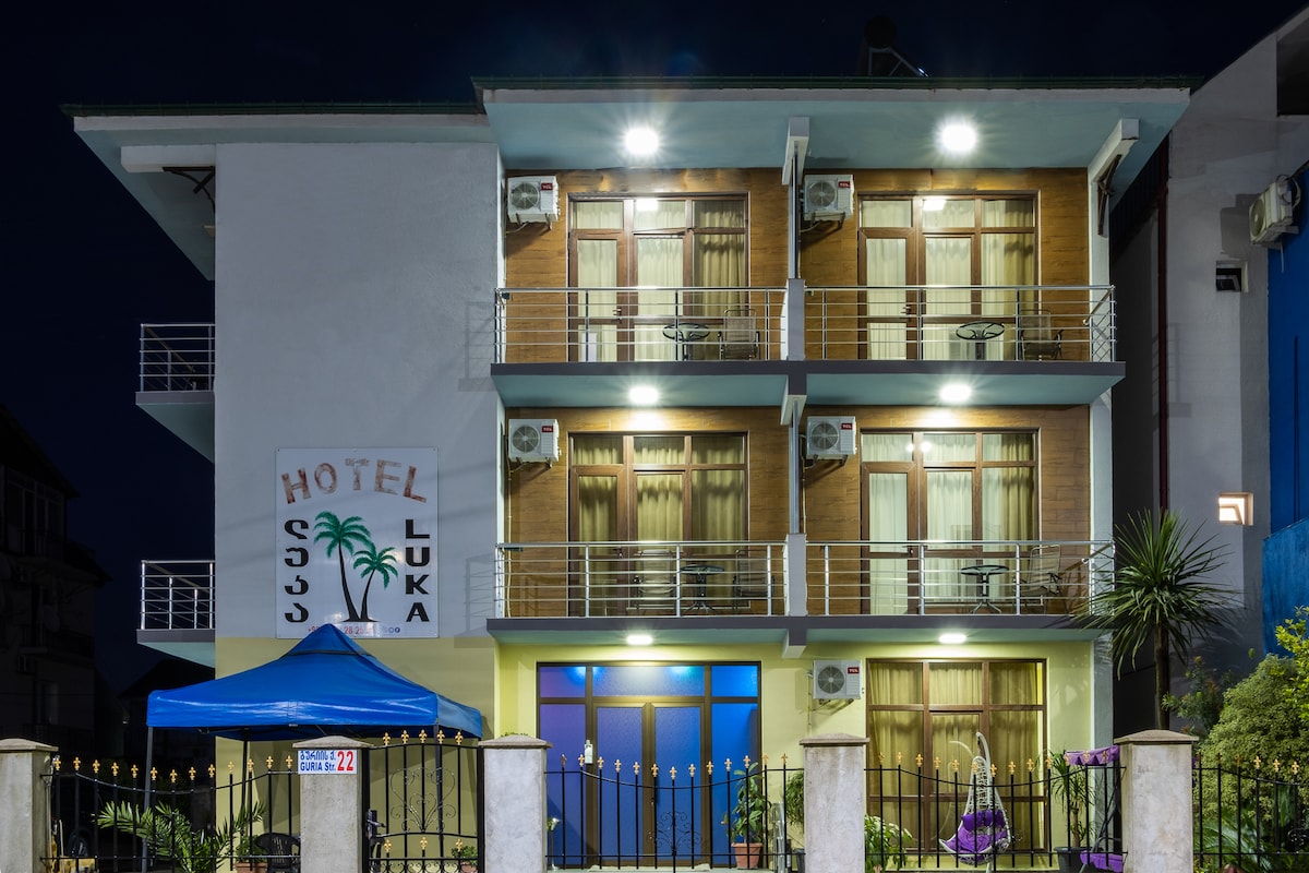 The hotel façade is illuminated at night, showcasing three levels of balconies with glass railings. Air conditioning units are visible under each balcony. A blue awning stands in front, providing shade to the entrance, which features a large logo sign welcoming guests.
