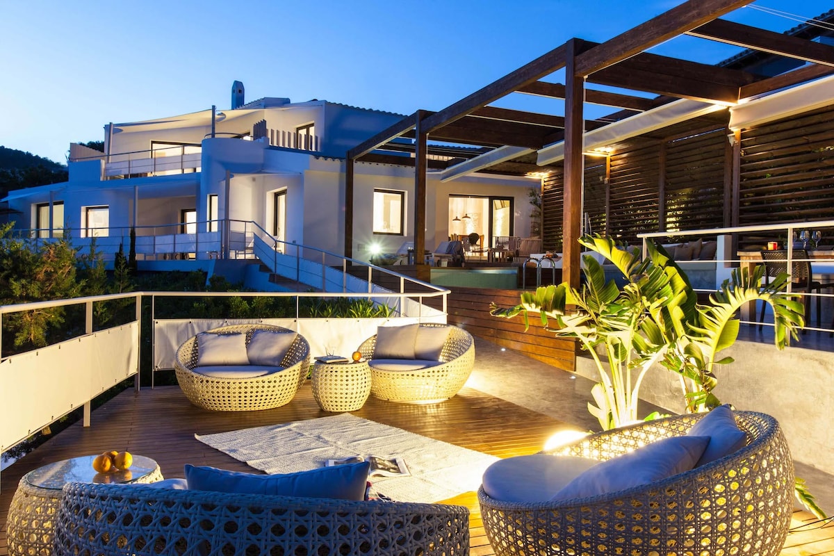 An outdoor terrace features comfortable seating with woven lounge chairs, surrounded by lush greenery. Soft lighting highlights the area, creating a relaxing ambiance. In the background, the modern villa stands against the evening sky, showcasing a blend of architecture and nature.