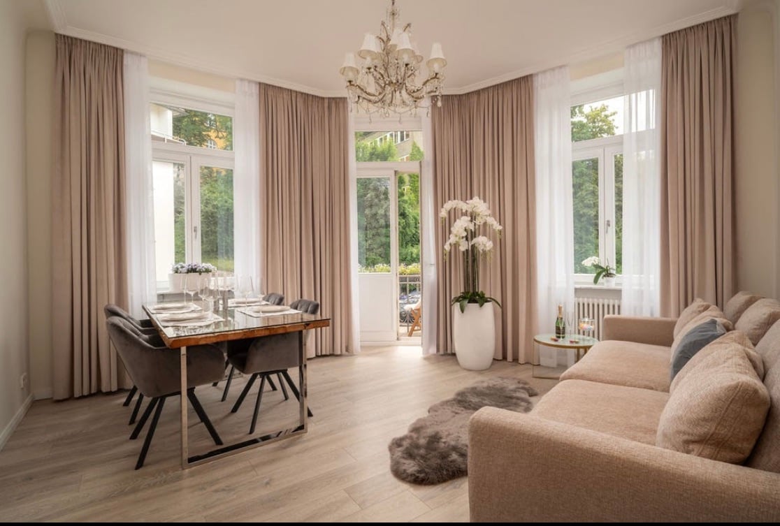 A spacious living and dining area features large windows adorned with soft drapes, allowing natural light to fill the room. A modern dining table is set for four, complemented by elegant chairs. A plush sofa is positioned nearby, with a stylish rug enhancing the floor.