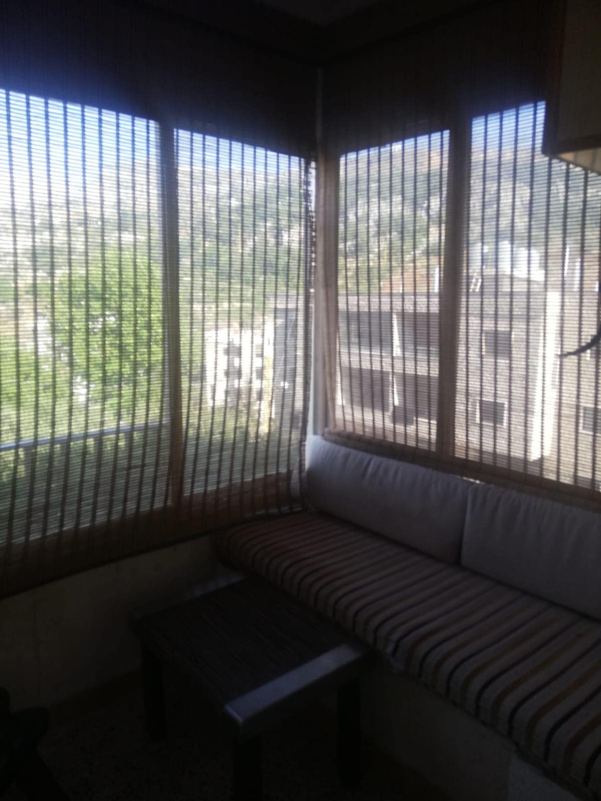 A comfortable seating area features a striped cushion along a wall of large windows, covered by bamboo blinds. Fresh greenery is visible outside, offering a serene view of the surrounding landscape.