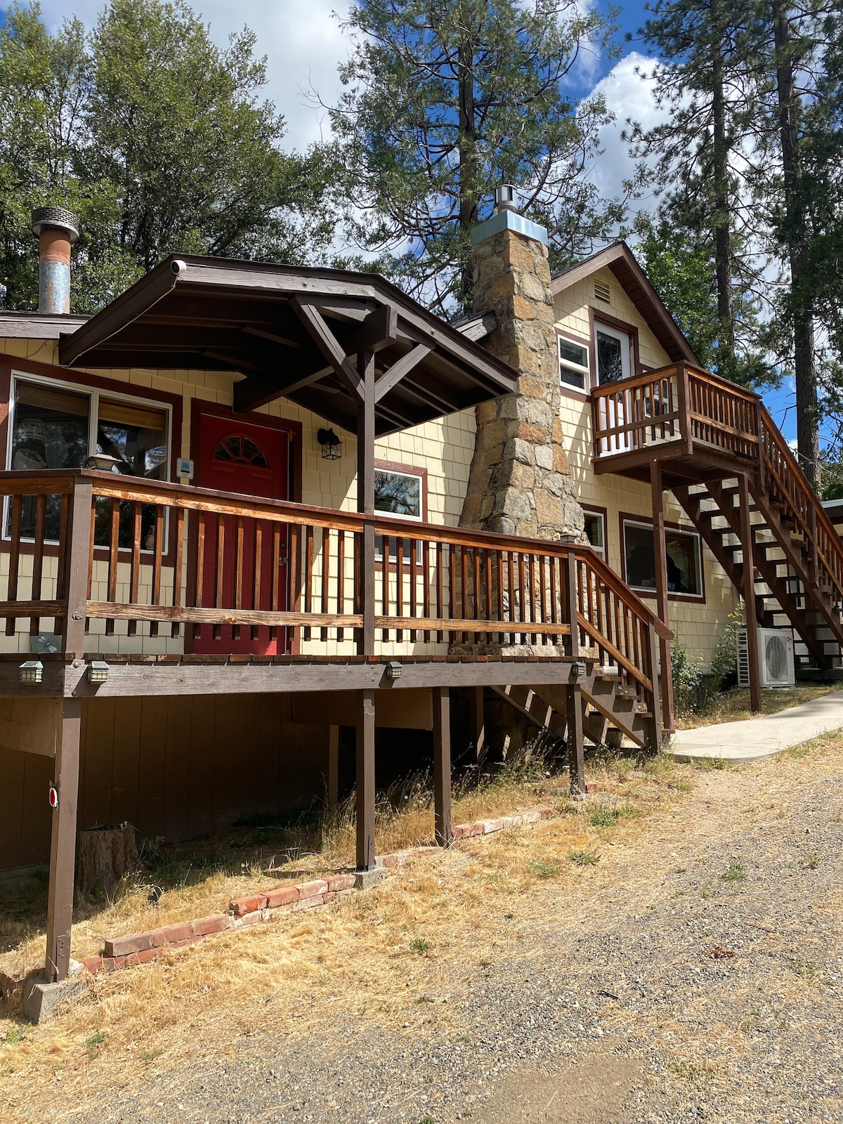 The exterior of the cabin features a welcoming entrance with a red door and a stone chimney. Wooden decks wrap around the structure, providing outdoor space. A staircase leads to the upper level, surrounded by trees and natural scenery.