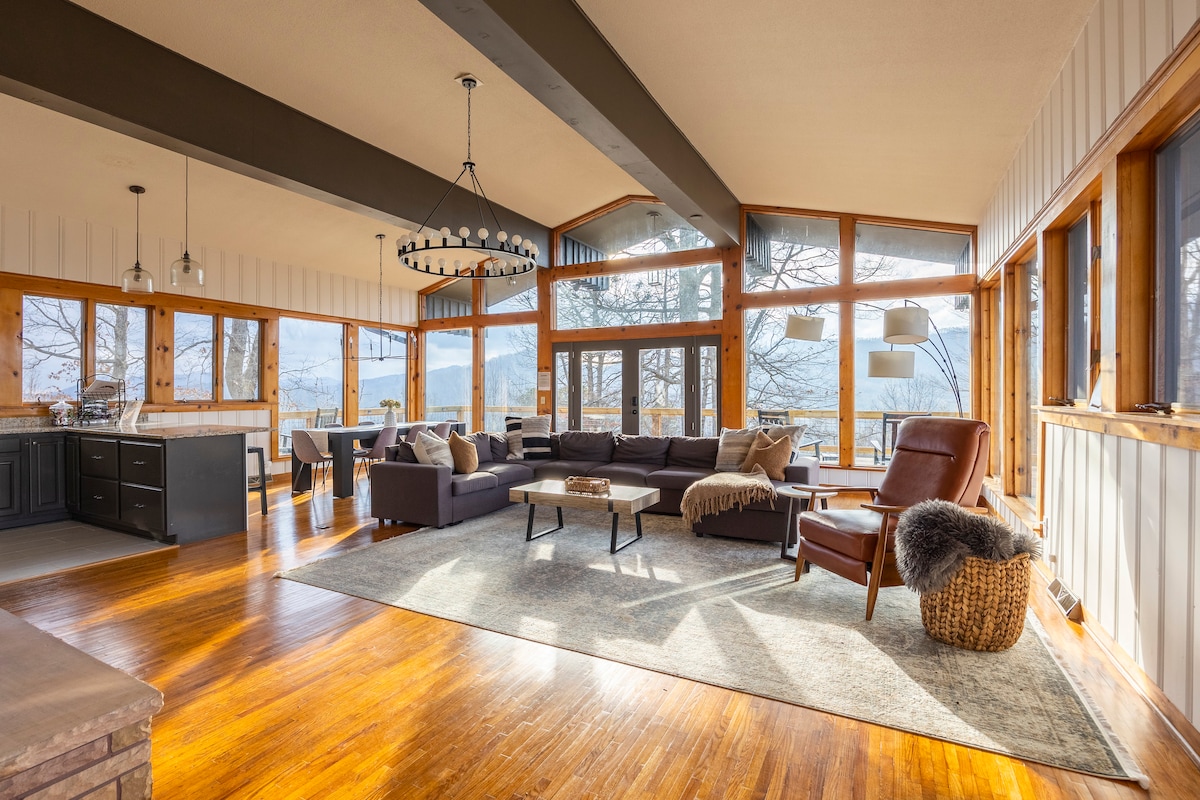 The spacious living area features abundant natural light from large windows overlooking the mountain views. A sectional sofa is arranged around a central coffee table on a soft area rug. A leather chair and cozy throw add to the inviting ambiance, with wood accents throughout.