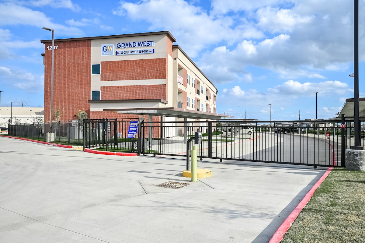 A modern, multi-story brick building with a prominent sign displaying 'Grand West' is set within a gated entrance. The paved driveway leads to a covered parking area, surrounded by landscaped green spaces and clear blue skies above.
