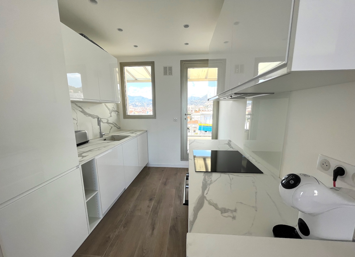 A modern kitchen with sleek white cabinetry is presented, featuring a minimalist design. Marble-patterned countertops and a contemporary stovetop are visible. Natural light floods the space through large windows, providing views of the surrounding hills.