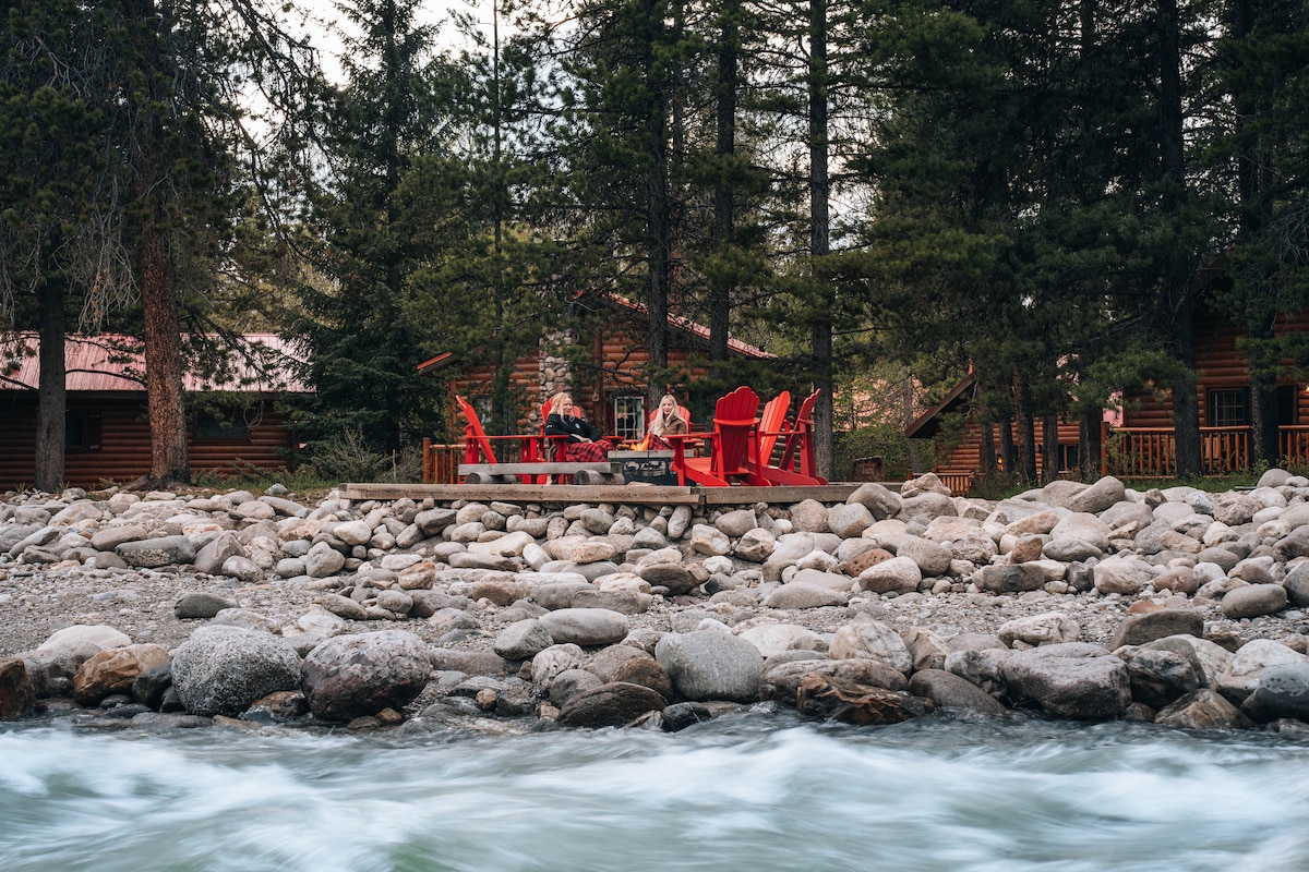 A wooden deck with red Adirondack chairs is positioned by a rocky riverbank. Tall evergreen trees surround the area, providing a natural backdrop. In the distance, rustic cabins are partially visible, set against a serene river flowing gently.