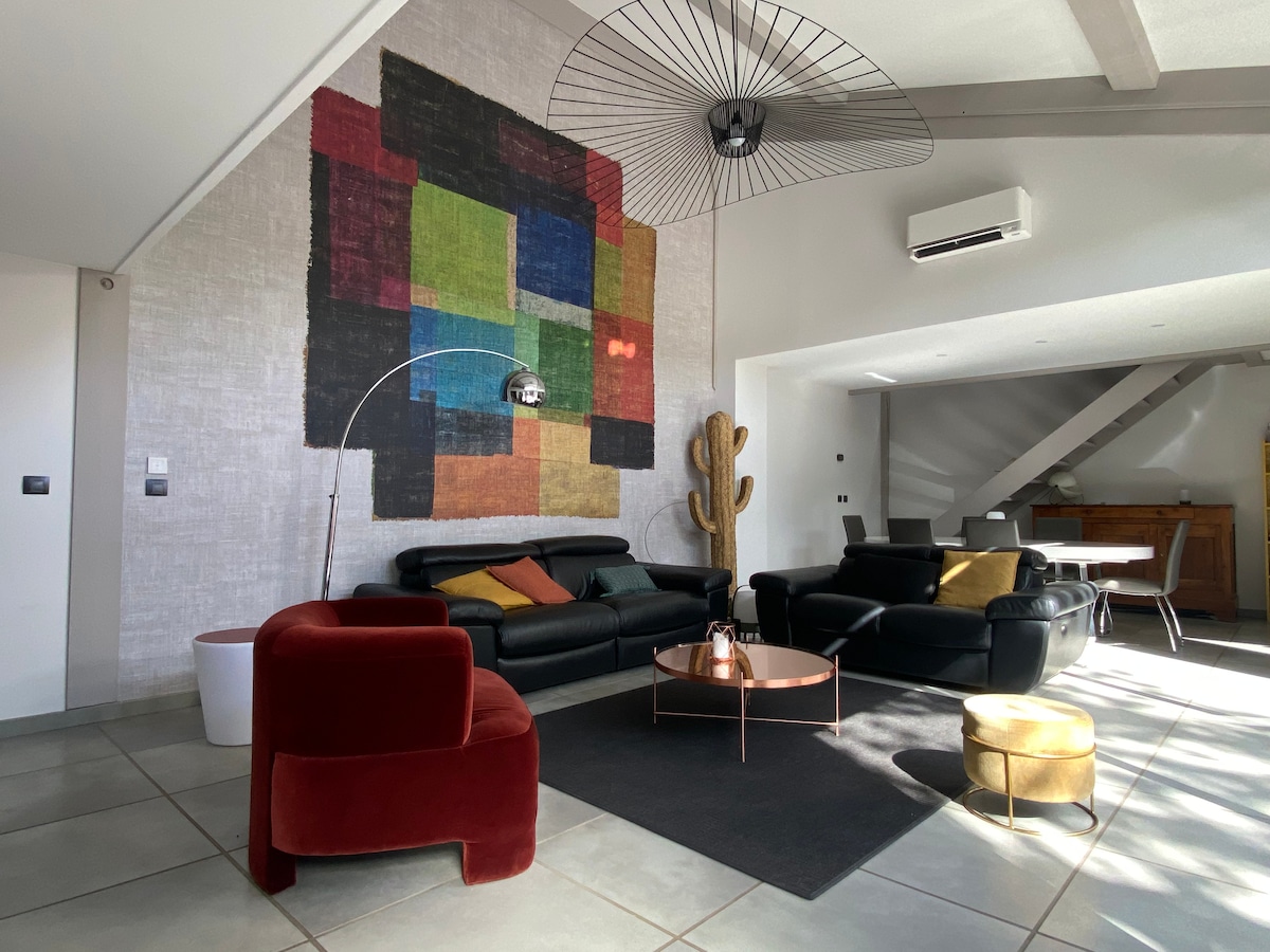 A spacious living area features a large mural in vibrant colors, complementing black leather sofas and a round coffee table. A cozy armchair in deep red adds a touch of warmth. Natural light fills the room through large windows, creating a bright atmosphere.