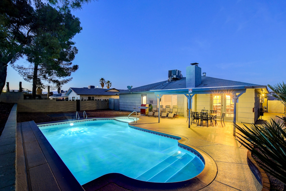 A spacious backyard is highlighted by a large swimming pool, shimmering under evening lights. Surrounding seating areas include lounge chairs and a dining table, positioned near the inviting pool. The exterior of the home, with its covered patio, is visible in the background.
