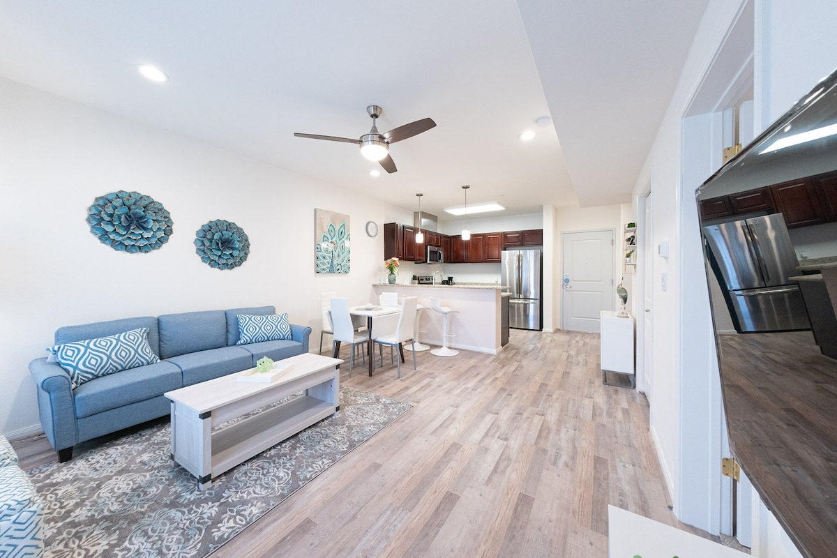 An open-concept living area features a blue sofa paired with a decorative area rug. A wooden coffee table sits in front, while the kitchen is visible in the background, showcasing dark wooden cabinets and stainless-steel appliances. Wall decor includes floral artwork.