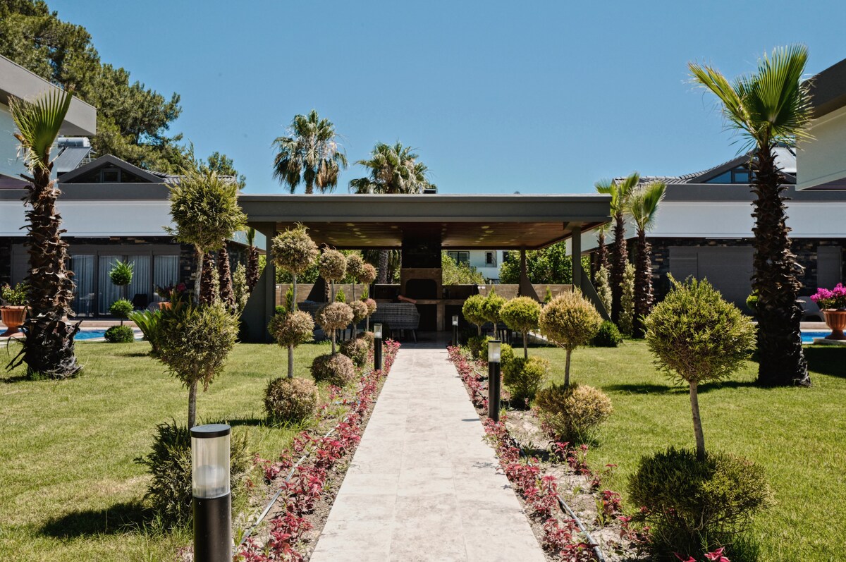 A stone pathway leads through a manicured garden featuring neatly trimmed shrubs and palm trees. A shaded seating area is visible at the end of the path, surrounded by lush greenery and vibrant flower beds, creating a serene outdoor space.