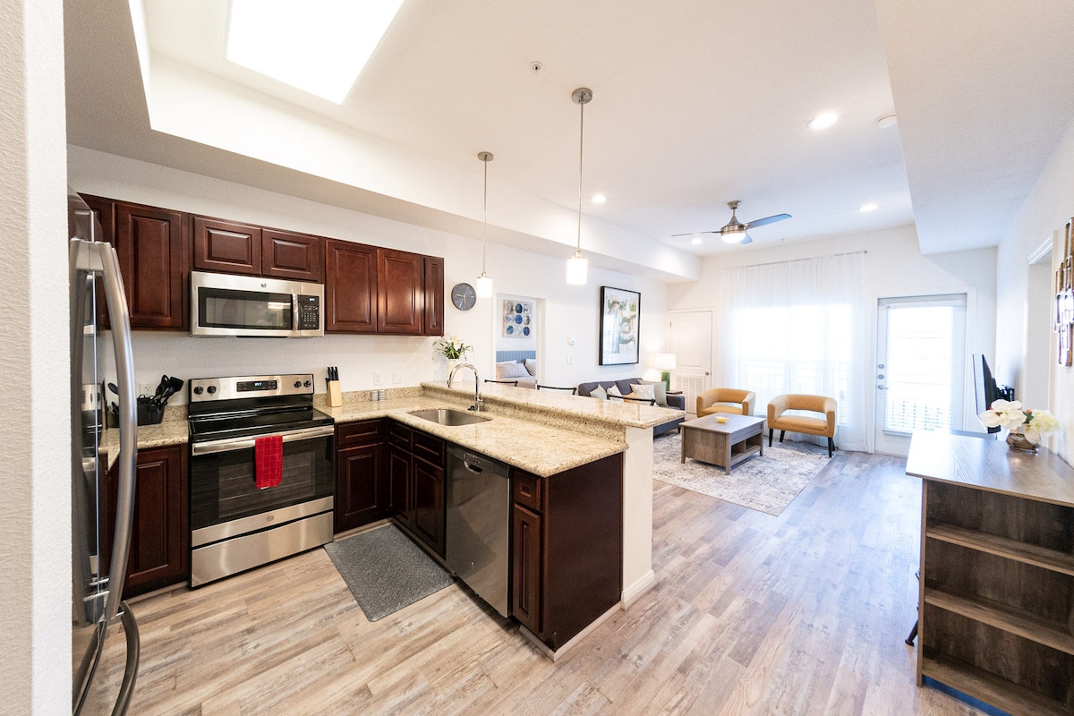 An open-concept kitchen and living area is displayed, featuring dark wood cabinetry and stainless steel appliances. A large island with granite countertops provides additional prep space, while natural light filters through large windows in the living area, which includes seating and decorative elements.