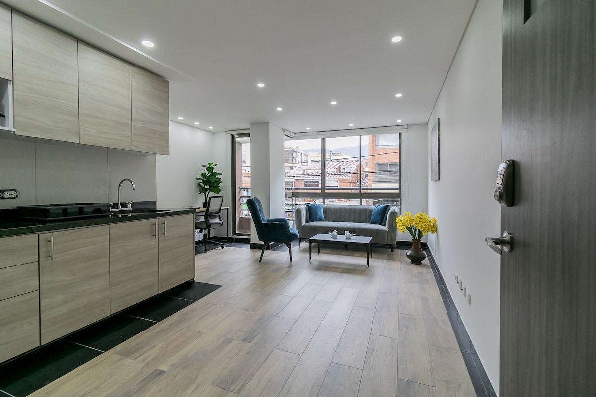An open living space features large windows allowing natural light to fill the area. A comfortable seating arrangement includes two armchairs and a sofa, complemented by a coffee table. Modern kitchen cabinetry is visible to the left, and indoor plants add a touch of greenery.