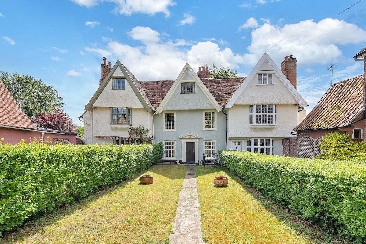 The exterior of the cottage displays a classic design, featuring a symmetrical façade with gabled roofs and crisp white walls. A manicured lawn leads to the entrance, framed by neatly trimmed hedges. The charming setting is complemented by a clear blue sky overhead.