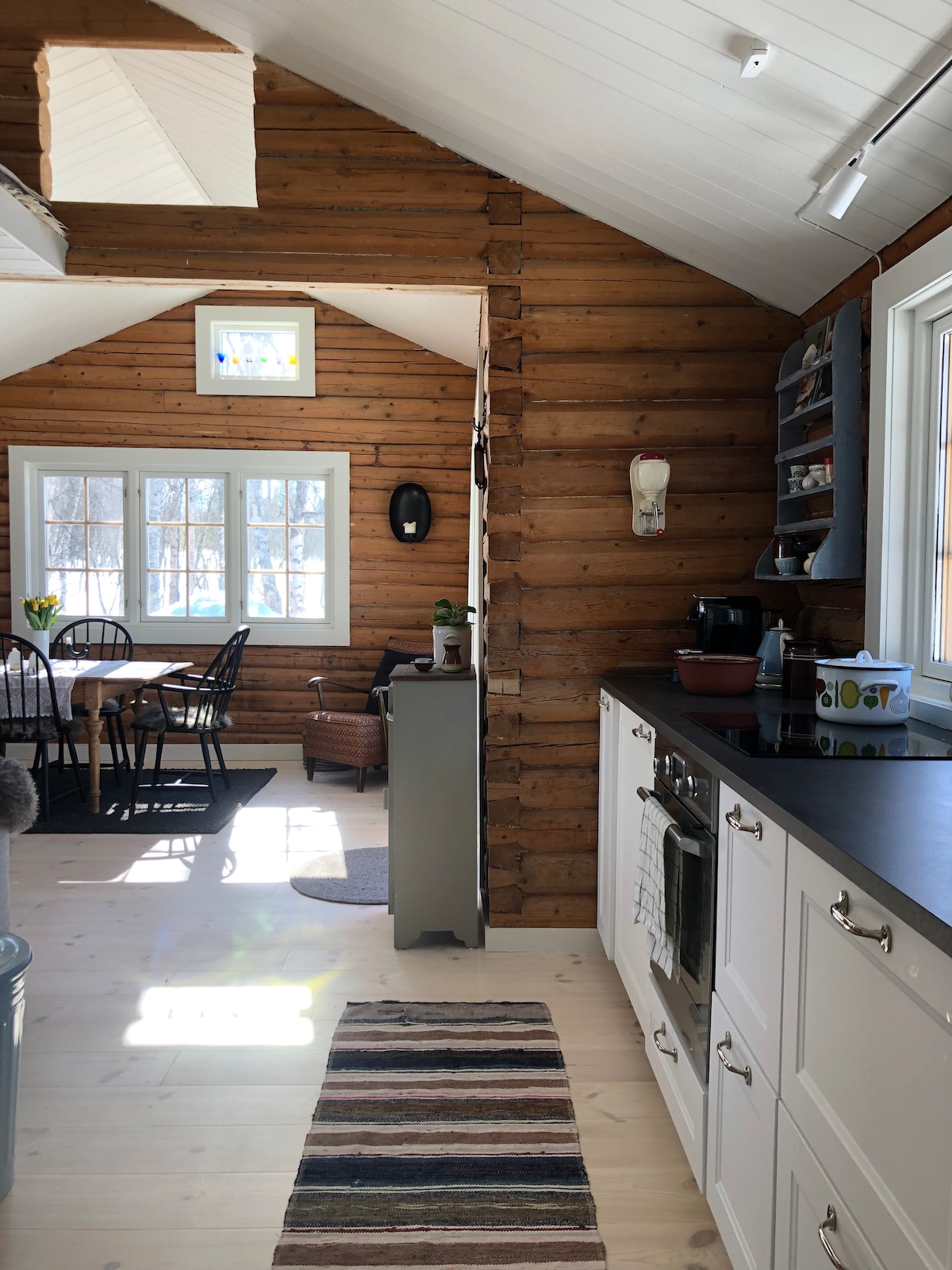 The kitchen area presents modern appliances and white cabinetry, contrasted by natural wooden walls. A compact dining space is visible with a round table and black chairs. Light filters through multiple windows, enhancing the brightness of the open floor plan.