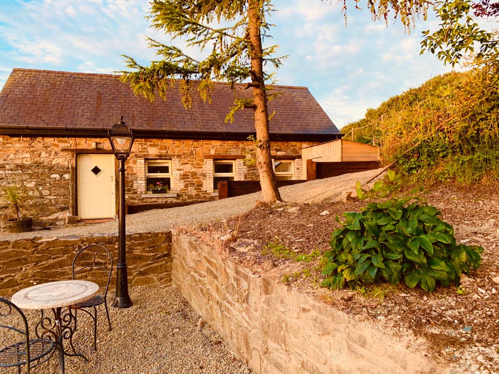 A charming stone cottage is seen amid natural surroundings. A small round table with two chairs is positioned on a gravel area beside a bush. A vintage-style lamp post stands nearby, and the cottage features a sloped roof under a clear sky.