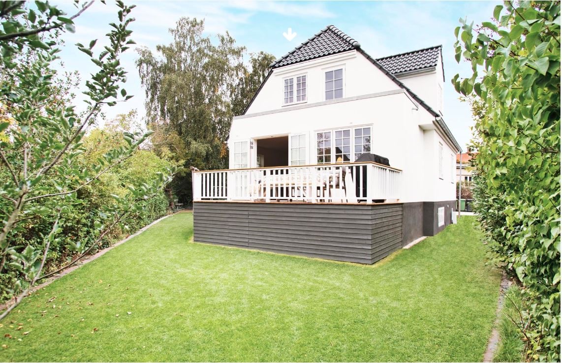 A modern villa is showcased with light-colored exterior walls and black accents. The spacious deck is furnished with a railing, providing outdoor seating options. Lush greenery surrounds the property, creating a serene environment in the yard.