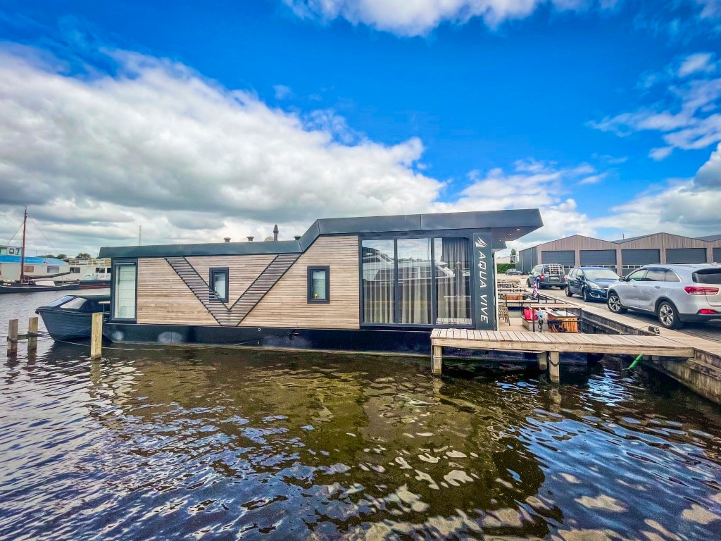 A modern houseboat is moored alongside a wooden dock, showcasing a sleek design with large windows that reflect the surrounding water. The exterior features a blend of wood and contemporary materials, with a clear blue sky above and clouds creating a serene atmosphere.
