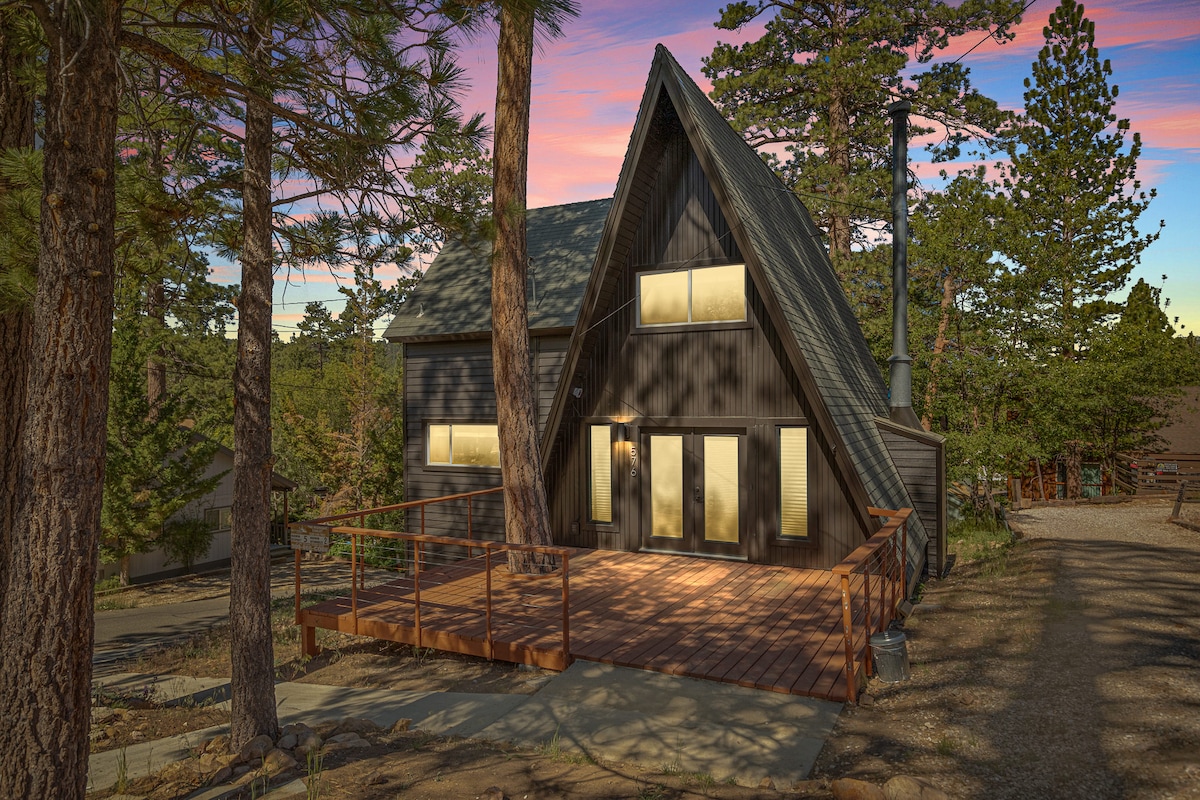 The modern A-Frame cabin is framed by tall pine trees, showcasing a spacious wooden deck. Large windows dominate the front facade, allowing natural light to illuminate the interiors. The gently sloped roof adds to the cabin's contemporary design, creating a welcoming outdoor space for relaxation.