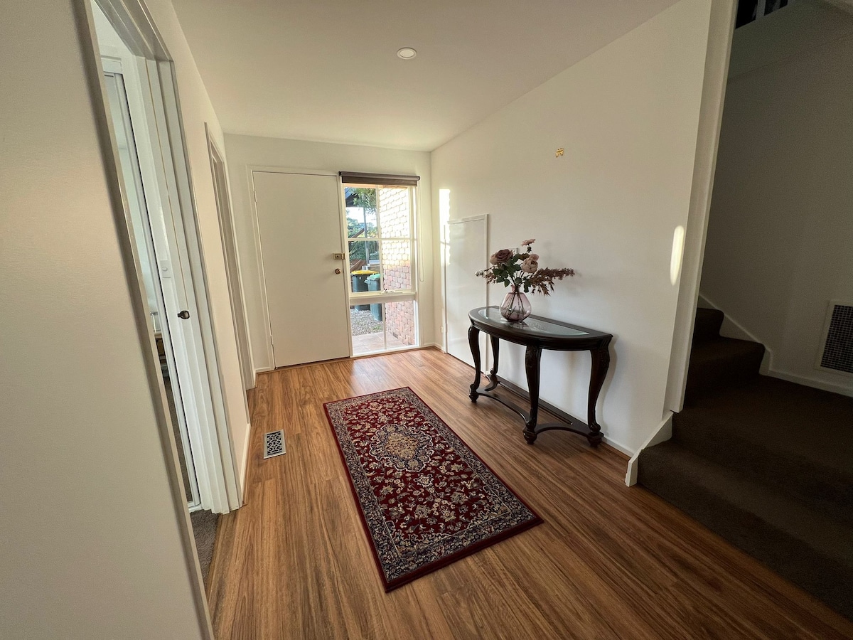 An entrance hall features wooden flooring and a decorative rug positioned along the center. A dark wooden console table sits against the wall, adorned with a vase containing fresh flowers. A doorway leads to an outdoor area, with natural light illuminating the space.