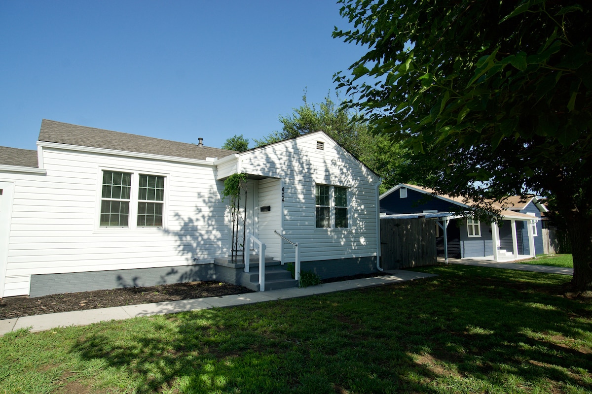 A single-story home is displayed, featuring white siding and several large windows. A small set of steps leads to the front door, while a green lawn and a narrow walkway enhance the exterior. Nearby, additional homes are visible under a clear blue sky.