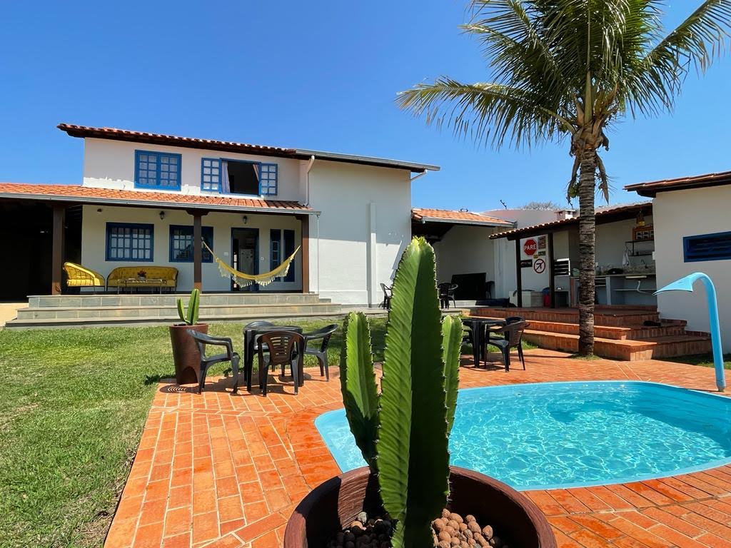 The image shows a charming two-story house with a red-tiled roof and a bright blue sky above. A small pool sits in the foreground, surrounded by a terracotta patio. A green cactus and palm tree provide a tropical touch, while seating areas are visible in the background.