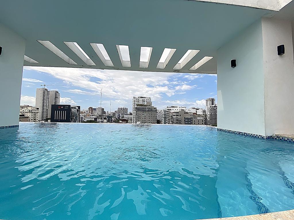 An outdoor pool area is outlined by an open structure, providing a view of the city skyline. The water appears calm and inviting, while the bright sky is adorned with scattered clouds, enhancing the serene atmosphere around the pool.