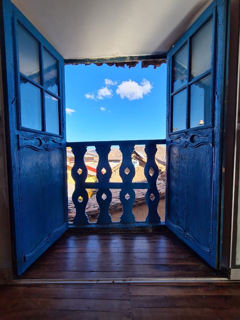 Two blue wooden doors are opened wide, revealing a view of a clear blue sky with scattered clouds. The rustic balcony railing is visible, enhancing the connection between indoor and outdoor spaces, while warm wooden flooring is seen inside the loft.
