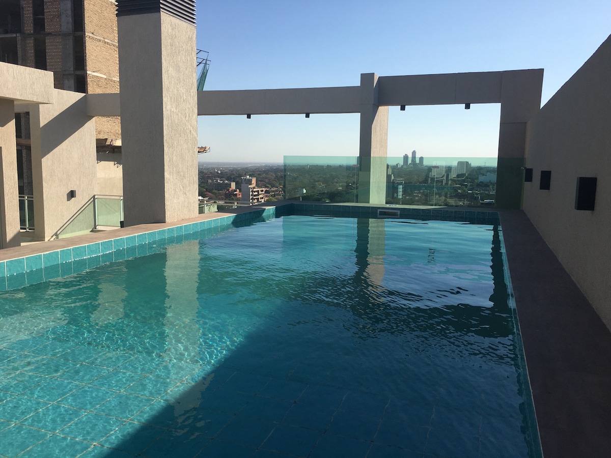 An outdoor pool area is visible atop a building, featuring clear blue water that reflects the sky. Surrounding glass railing enhances the panoramic view of the city skyline, while the pool deck is tiled in light blue, providing a visually appealing contrast.