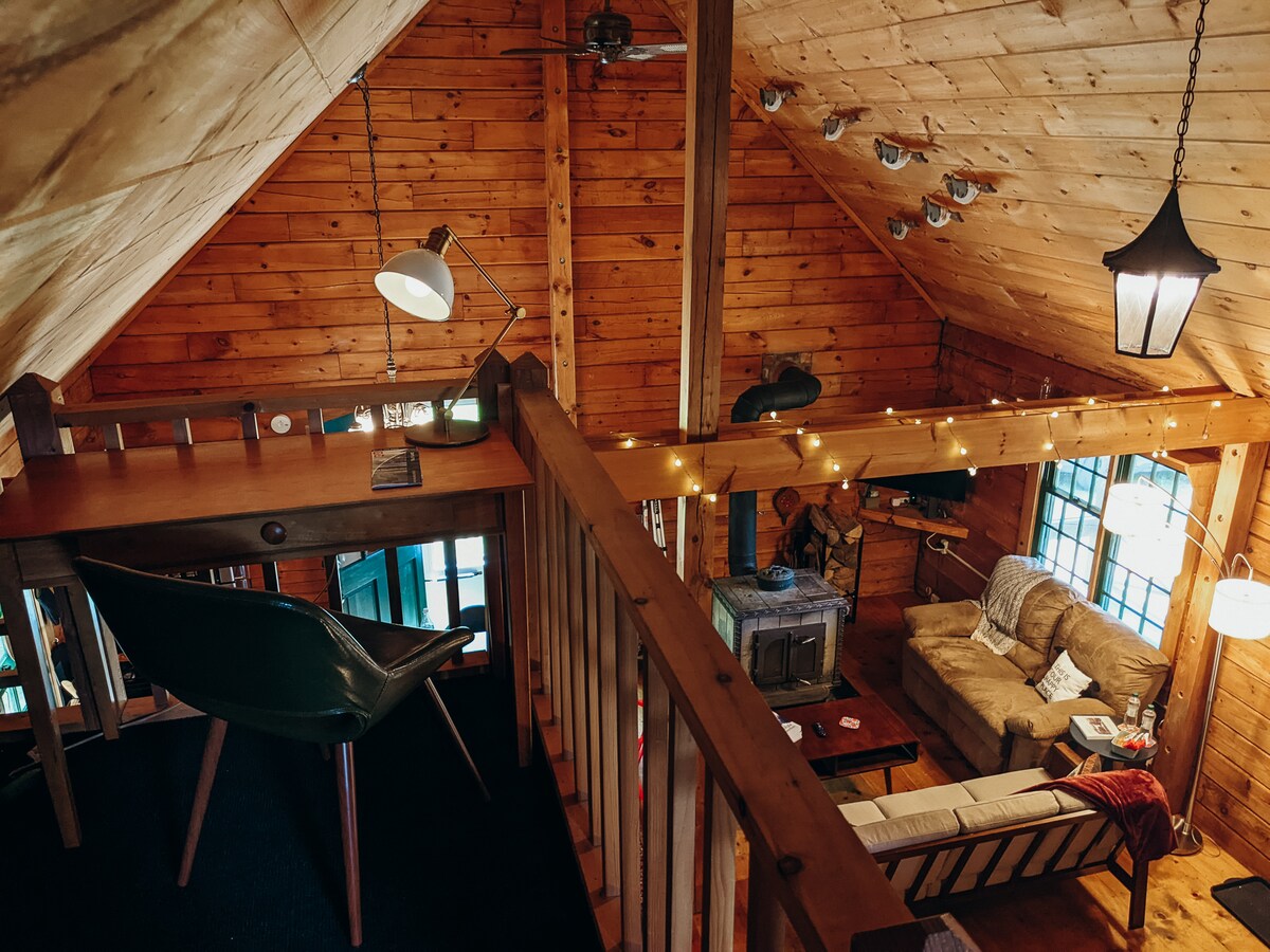 A loft area overlooks the welcoming living space below, featuring a writing desk and a comfortable chair. The warm wood paneling creates an inviting ambiance. Natural light filters through the windows, highlighting the cozy seating arrangement and decor in the living area.