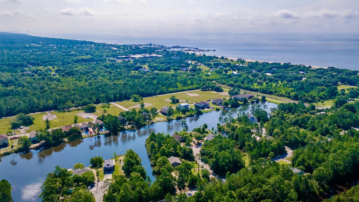An aerial view showcases a serene landscape, featuring lush greenery surrounding a calm lake. In the distance, the coastline and beach are visible, blending with the tranquil environment. The scene captures a peaceful community set against a clear sky.