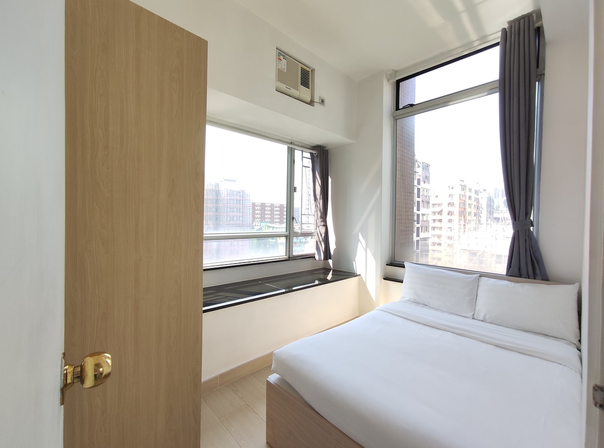 A well-lit bedroom features large windows that provide ample natural light and a view of the surrounding area. A spacious bed is dressed in crisp white linens, and a wooden door leads to the room. Light-colored walls complement the minimalist design.