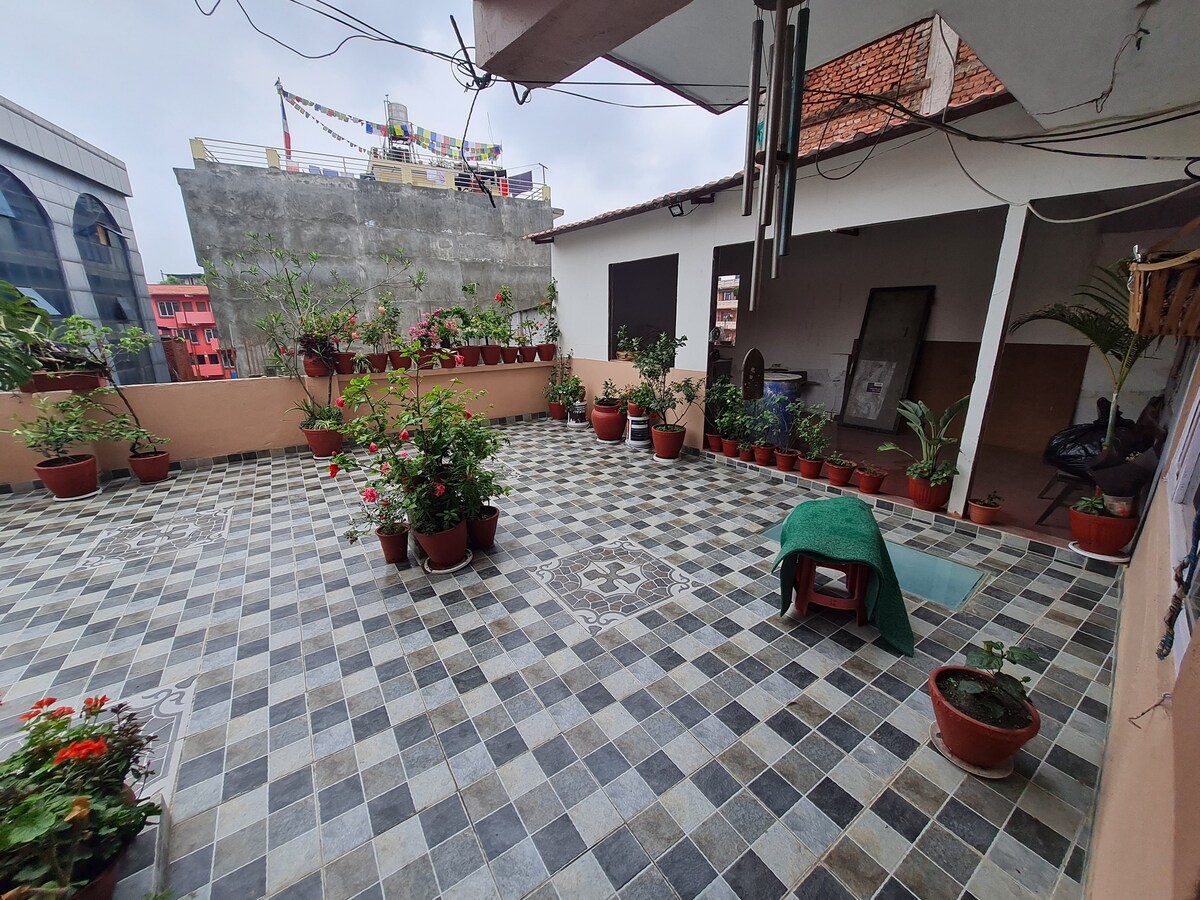 A spacious terrace features a tiled floor adorned with decorative patterns. Potted plants, including colorful flowers, are arranged throughout the area, creating a vibrant setting. A green mat is visible alongside a chair, offering a comfortable spot to relax.