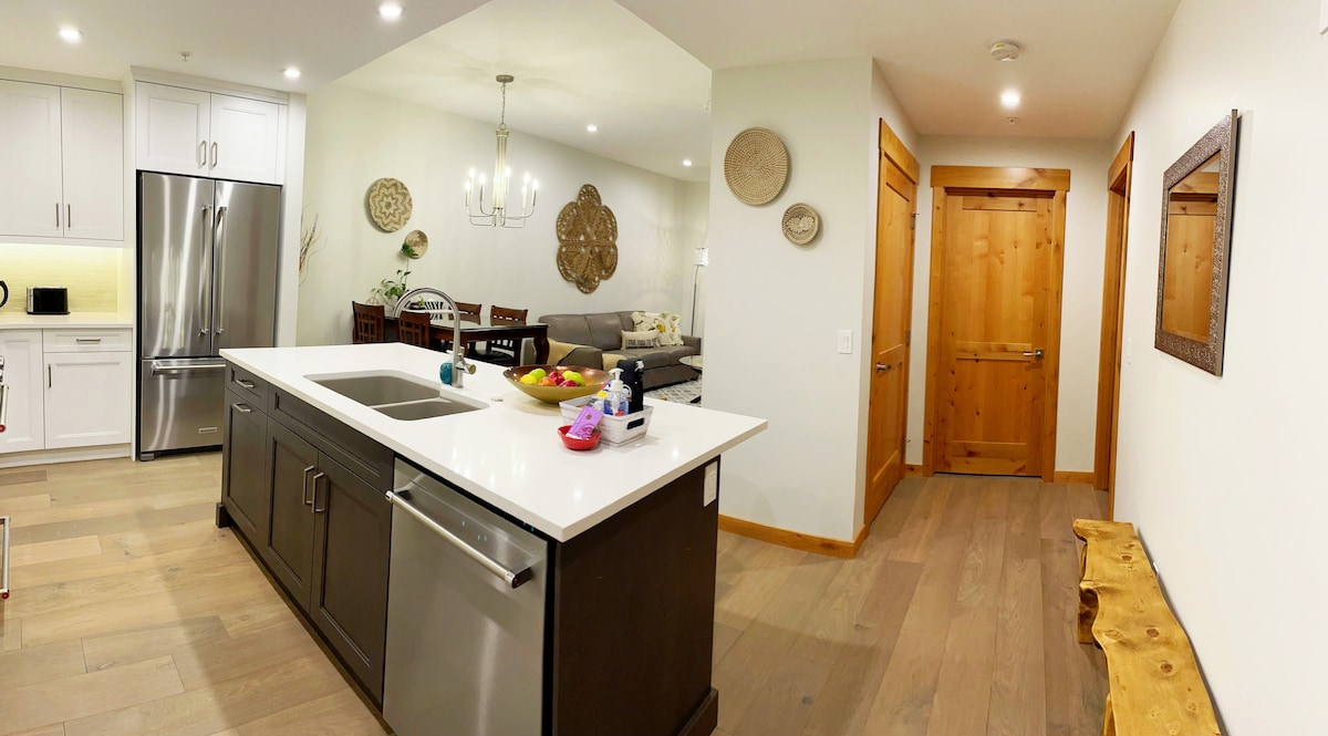 An open kitchen area features a large island with a sink and fruit, surrounded by modern stainless-steel appliances. A dining space with wooden chairs is visible in the background. Natural light brightens the room, while warm wood accents and decor add a cozy touch.