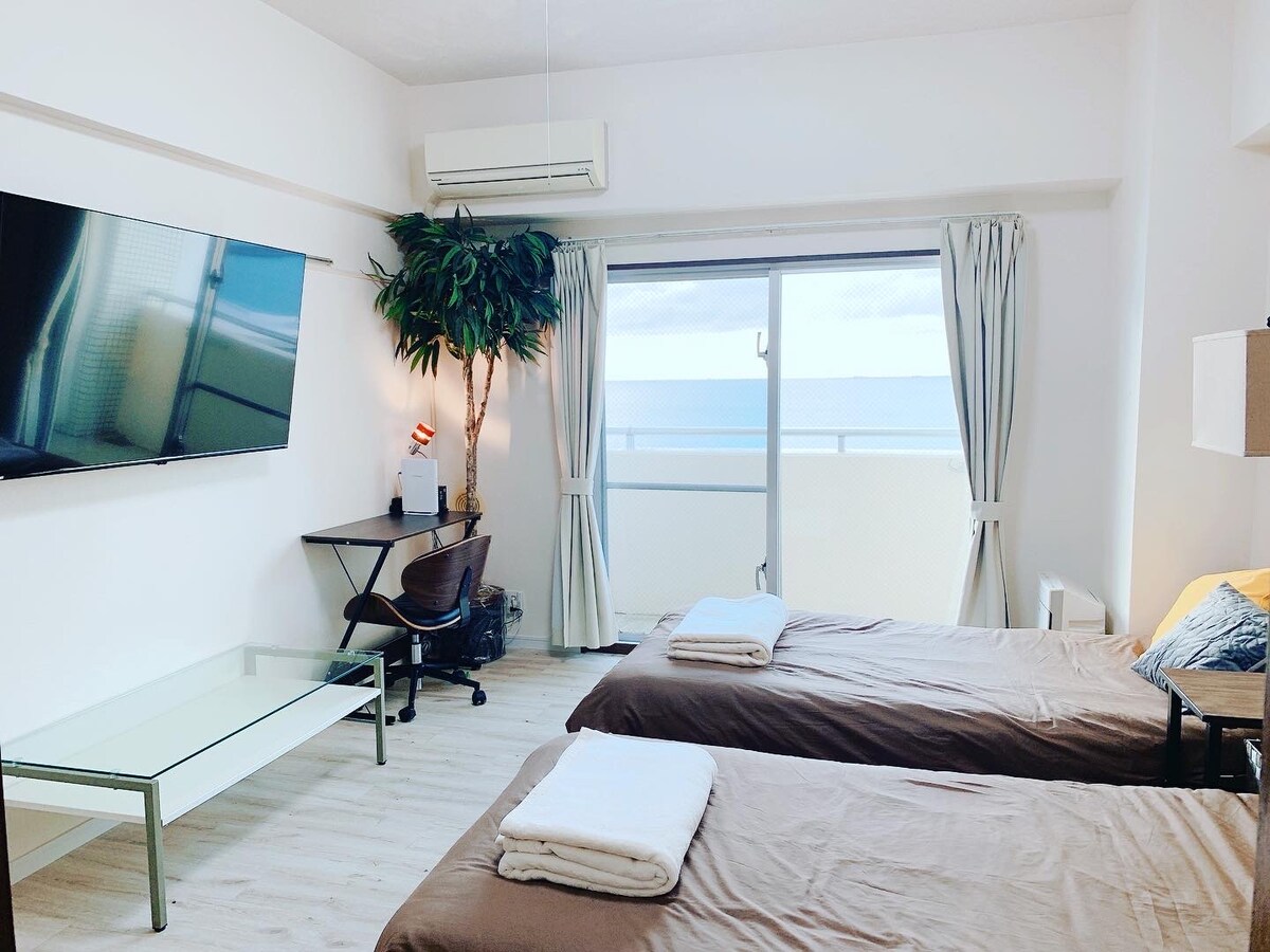 A bright room features two spacious beds with neutral bedding, facing a large window that provides ocean views. A flat-screen television is mounted on the wall, and a modern desk with a rolling chair sits in the corner. Light and airy curtains frame the window.